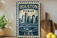 A vintage-style metal sign with the text "HOUSTON TEXAS U.S.A." displayed prominently, featuring a cityscape illustration. The sign is placed on a wooden surface, with a potted plant, a wooden cutting board, and some lemons nearby.