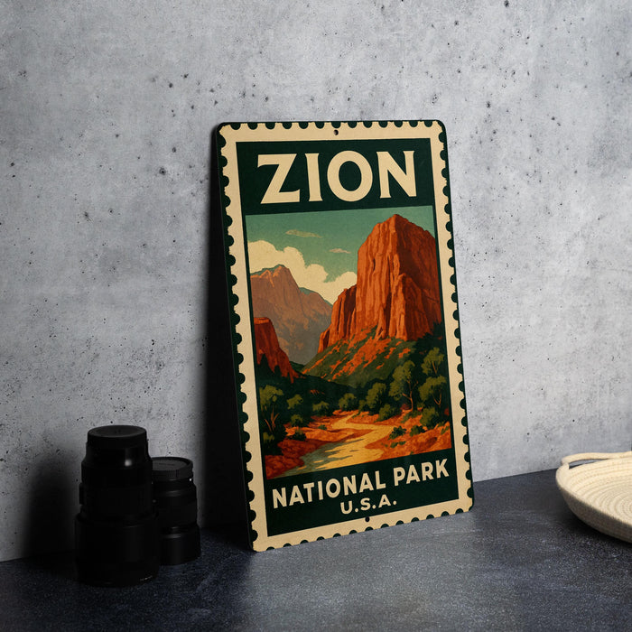 A vintage-style metal sign for Zion National Park, featuring a mountain landscape with a river and trees, is displayed on a gray concrete surface.