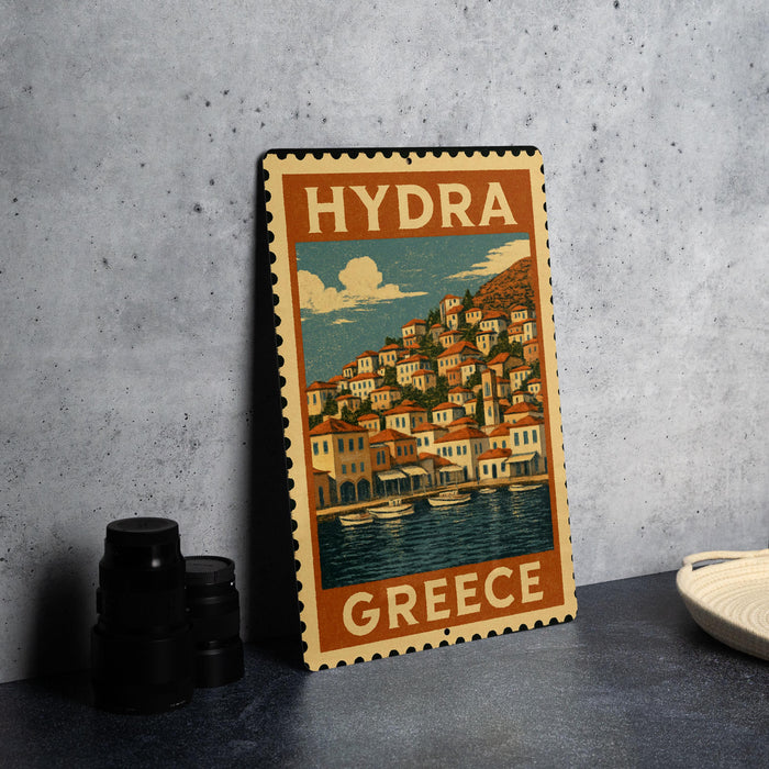 A vintage-style metal stamp depicting a coastal town in Greece, with a cloudy sky and water in the background.