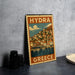 A vintage-style metal stamp depicting a coastal town in Greece, with a cloudy sky and water in the background.