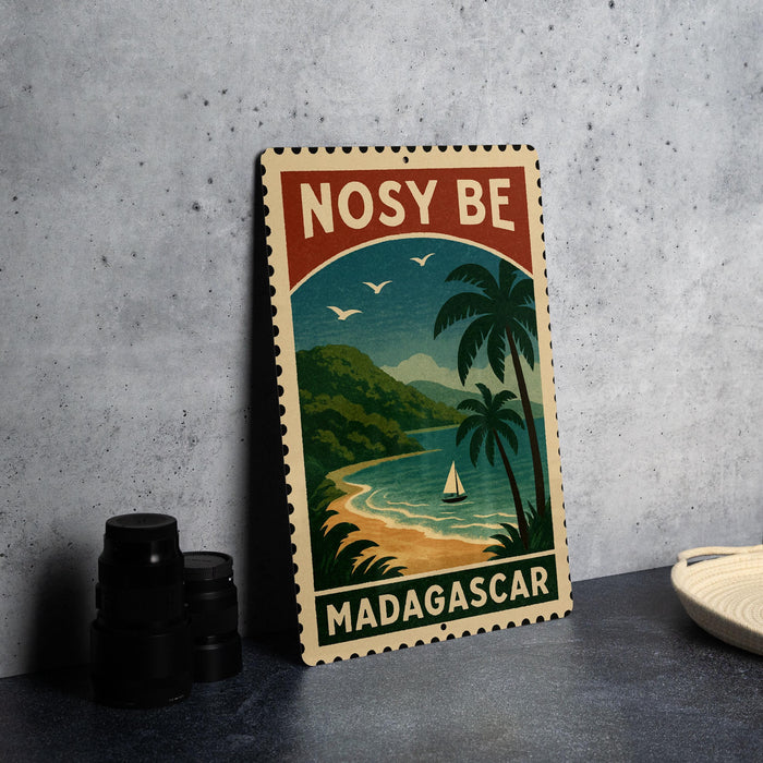A vintage-style metal sign with the text "NOSY BE MADGASCAR" and a tropical beach scene with palm trees, a sailboat, and a mountain backdrop.