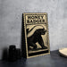 a metal sign with a black and white illustration of a honey badger, placed on a gray countertop next to some black camera lenses.