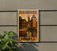 A vintage-style postage stamp depicting a cityscape of Strasbourg, France, with a castle and other buildings, hanging on a wall.