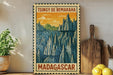 A vintage-style stamp depicting a mountainous landscape with the text "TSINGY DE BEMARAH" and "MADAGASCAR" printed on it, placed on a wooden surface next to a potted plant and a wooden cutting board.