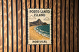 A vintage-style metal sign with the text "PORTO SANTO ISLAND" and an illustration of a beach and mountains, mounted on a wooden wall.