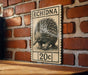 a vintage-style stamp or postcard with an illustration of an echidna, a type of small insect, on it. The stamp is mounted on a brick wall, and there is a wooden table below it.