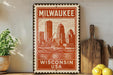 A vintage-style metal sign with the text "Milwaukee, Wisconsin, USA" printed on it, featuring an illustration of the Milwaukee skyline. The sign is displayed on a shelf next to a wooden cutting board and a potted plant.
