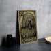 A wooden postcard featuring a black and white illustration of a shaggy dog's head, with the text "KOMONDOR" printed on it. The postcard is placed on a table next to some black camera lenses.