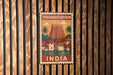 A vintage postage stamp from India featuring a temple surrounded by palm trees and buildings, hanging on a wooden wall.