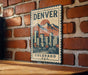 A vintage-style metal sign with the text "DENVER COLORADO U.S.A." displayed on a brick wall.