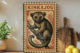 A vintage-style metal sign featuring a cartoon illustration of a kangaroo, with the text "KINKAJOU" prominently displayed. The sign is placed on a wooden surface, with a potted plant, a wooden cutting board, and some lemons nearby.