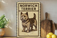 a decorative sign with a picture of a dog, specifically a Norwich Terrier, on it. The sign is placed on a wooden surface, surrounded by various kitchen utensils and items, such as a wooden cutting board, a knife, a spoon, and a bowl.