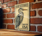 A vintage postage stamp featuring a great blue heron, mounted on a brick wall.