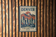 A vintage-style metal sign with the word "DENVER" and a mountain landscape, mounted on a wooden wall.
