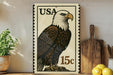 A framed stamp-like artwork depicting a bald eagle, with the text "USA 15c" printed on it, is displayed on a shelf next to a wooden cutting board and some kitchen utensils.
