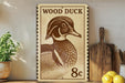 a wooden duck stamp with the text "WOOD DUCK 8c" on it, placed on a wooden surface next to a potted plant and a wooden cutting board.