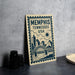 A framed poster of Memphis, Tennessee, USA, featuring a cityscape and a bridge, is displayed on a table next to some black camera lenses.
