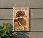 A wooden duck stamp is mounted on a wall, with a plant visible to the left.
