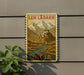 A vintage-style sign depicting a mountainous landscape with a small village or settlement, mounted on a wall.