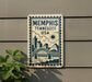 A sign that reads "Memphis, Tennessee, USA" with a cityscape illustration, mounted on a wall next to a plant.