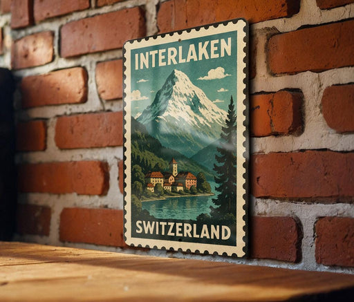 A vintage-style metal stamp depicting a mountain landscape with a village in the foreground, hanging on a brick wall.
