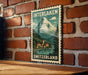 A vintage-style metal stamp depicting a mountain landscape with a village in the foreground, hanging on a brick wall.
