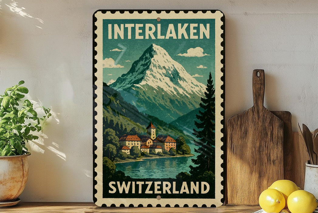 A vintage-style metal sign with a mountain landscape and Swiss village, featuring the text "INTERLAKEN SWITZERLAND" and a postage stamp-like border.