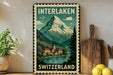 A vintage-style metal sign with a mountain landscape and Swiss village, featuring the text "INTERLAKEN SWITZERLAND" and a postage stamp-like border.