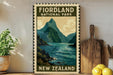 a vintage-style poster of Fiordland National Park in New Zealand, with a mountain landscape and a body of water. The poster is displayed on a wall, and there are some kitchen utensils, such as a cutting board and a knife, as well as some potted plants and lemons in the foreground.
