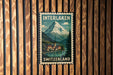 A vintage-style metal sign with the text "INTERLAKEN SWITZERLAND" and an illustration of a mountain landscape with a village in the background.