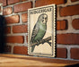 A vintage-style postage stamp featuring a parakeet, mounted on a brick wall.