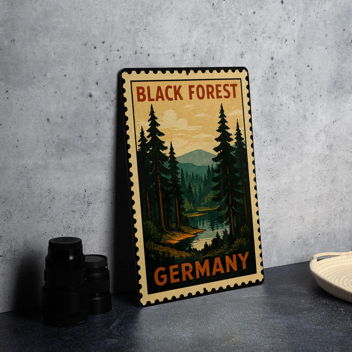 A vintage-style metal stamp depicting a forest scene with pine trees and a river, placed on a gray countertop next to some black camera lenses.
