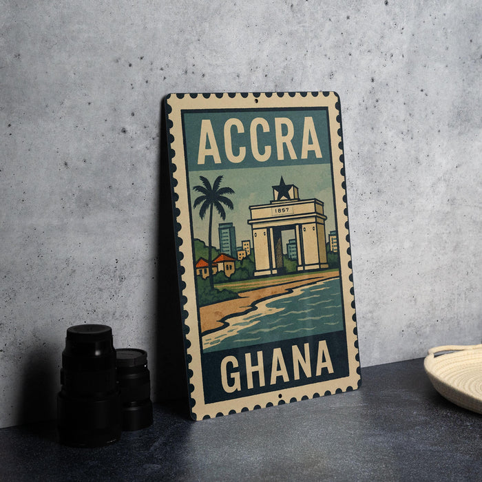 A metal sign with the text "ACCRA GHANA" and an illustration of a cityscape with a palm tree, placed on a gray countertop next to some black camera lenses.
