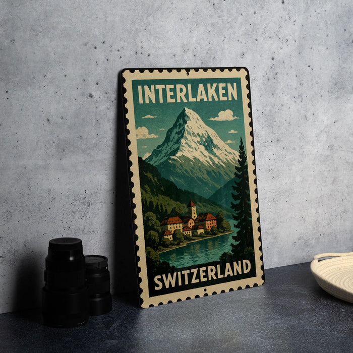 A vintage-style metal sign with a mountain landscape and a Swiss village, featuring the text "INTERLAKEN SWITZERLAND".