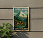 A vintage-style postage stamp depicting a mountain landscape with a village in the foreground, hanging on a wall.