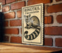 a sign with a raccoon illustration on it, mounted on a brick wall.