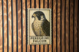 A postage stamp featuring a peregrine falcon is displayed against a wooden background.
