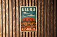 A vintage-style poster of Uluru, a famous rock formation in Australia, is displayed on a wooden wall.