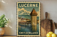 A vintage-style poster of Lucerne, Switzerland, featuring a castle and a bridge over a river, with mountains in the background. The poster is framed and displayed on a wall, with a wooden cutting board and a potted plant to the left.