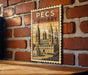 A vintage-style poster of the Pecs Cathedral in Hungary, featuring a castle-like structure with multiple towers and spires, is displayed on a brick wall.
