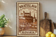 A vintage-style stamp or sign depicting the Bratislava Castle in Slovakia, with a wooden cutting board and a potted plant in the foreground.