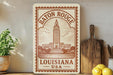 A vintage-style metal sign with the text "Baton Rouge, Louisiana, USA" printed on it, featuring an illustration of the Louisiana State Capitol building.