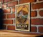 A vintage-style metal sign with a landscape illustration and the text "SICILY ITALY" is displayed on a brick wall.