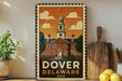 A framed vintage-style poster of Dover, Delaware, featuring a building with a steeple and a clock tower, is displayed on a shelf next to a wooden cutting board and some potted plants.