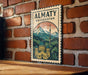 A vintage-style poster of Almaty, Kazakhstan, featuring a mountainous landscape with a domed building and a tower, mounted on a brick wall.
