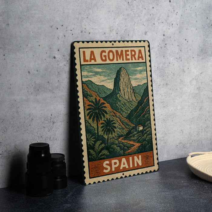 A vintage metal sign with a landscape illustration and the text "LA GOMERA" and "SPAIN" printed on it, placed on a gray countertop next to some black camera lenses.
