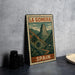 A vintage metal sign with a landscape illustration and the text "LA GOMERA" and "SPAIN" printed on it, placed on a gray countertop next to some black camera lenses.