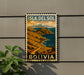 A vintage-style postage stamp depicting a mountainous landscape with a river, hanging on a wall next to a plant.