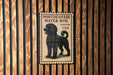 A vintage-style postage stamp featuring a black poodle, mounted on a wooden wall.