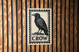 a black and white stamp-like sign with a black crow illustration on a wooden background.